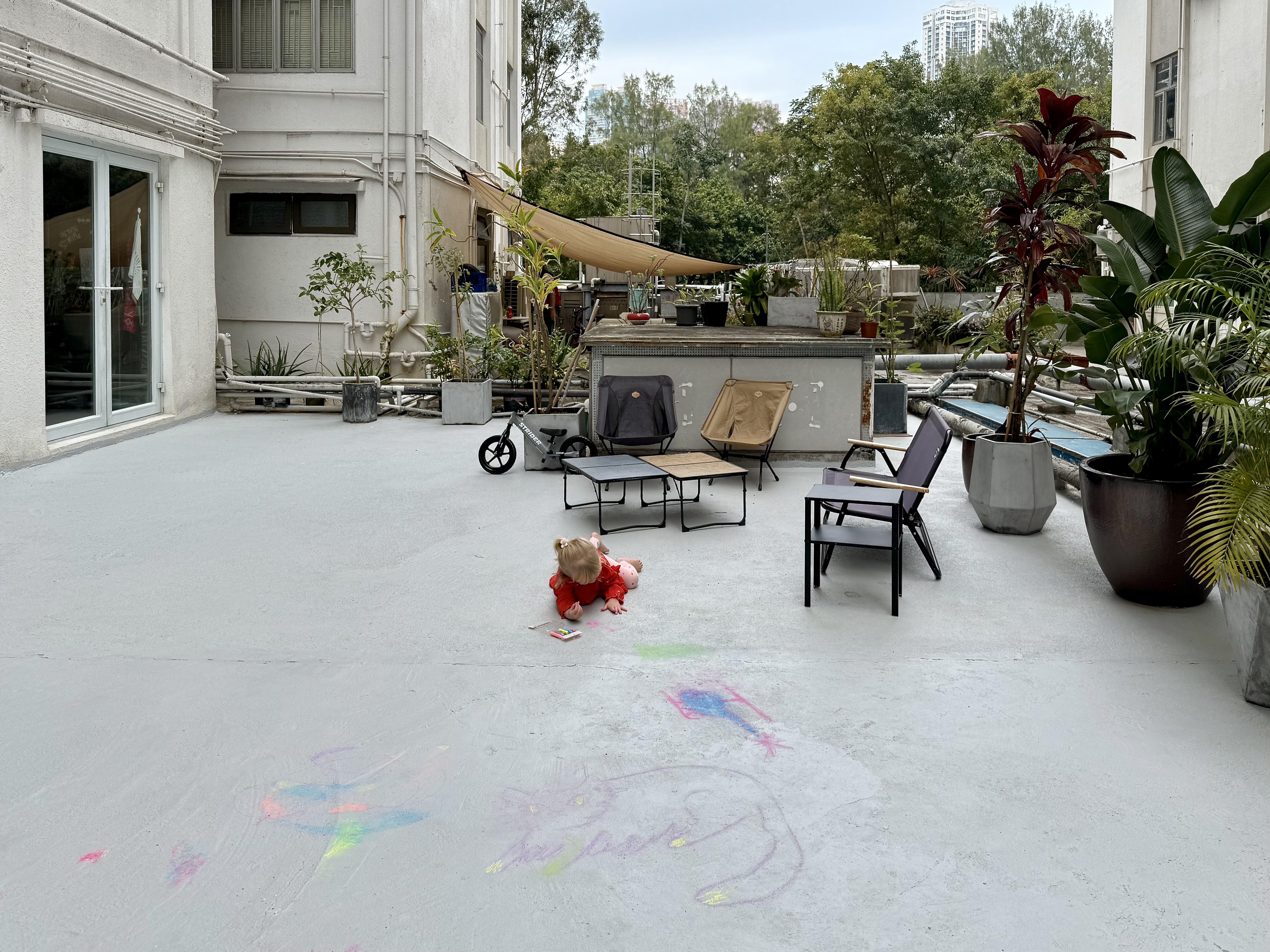 A girl drawing with chalk on an outdoor terrace, with chairs and a bike behind her. Tropical plants and trees frame the buildings.