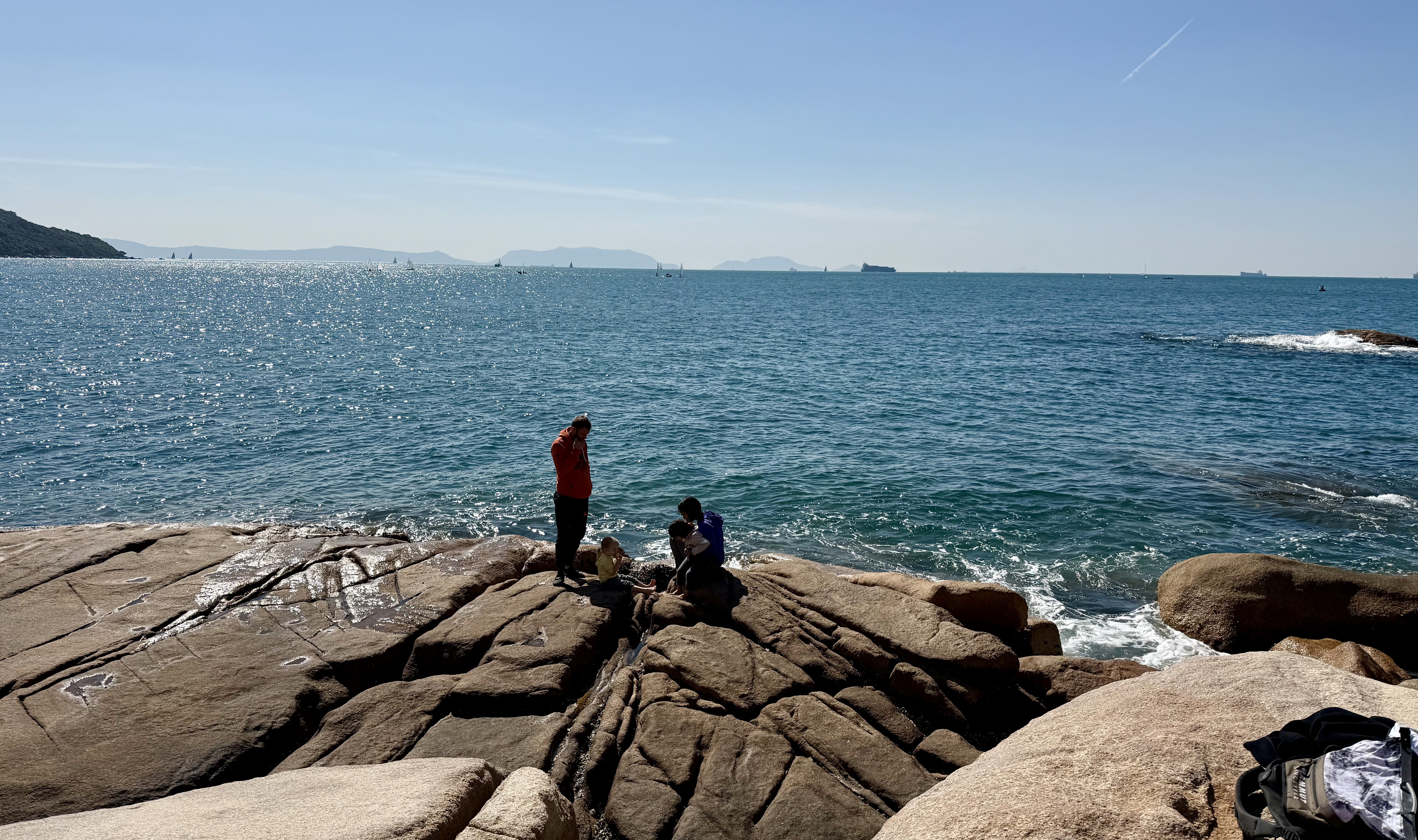 Children play on the rocks with sailboats, the ocean, and container ships visible behind.